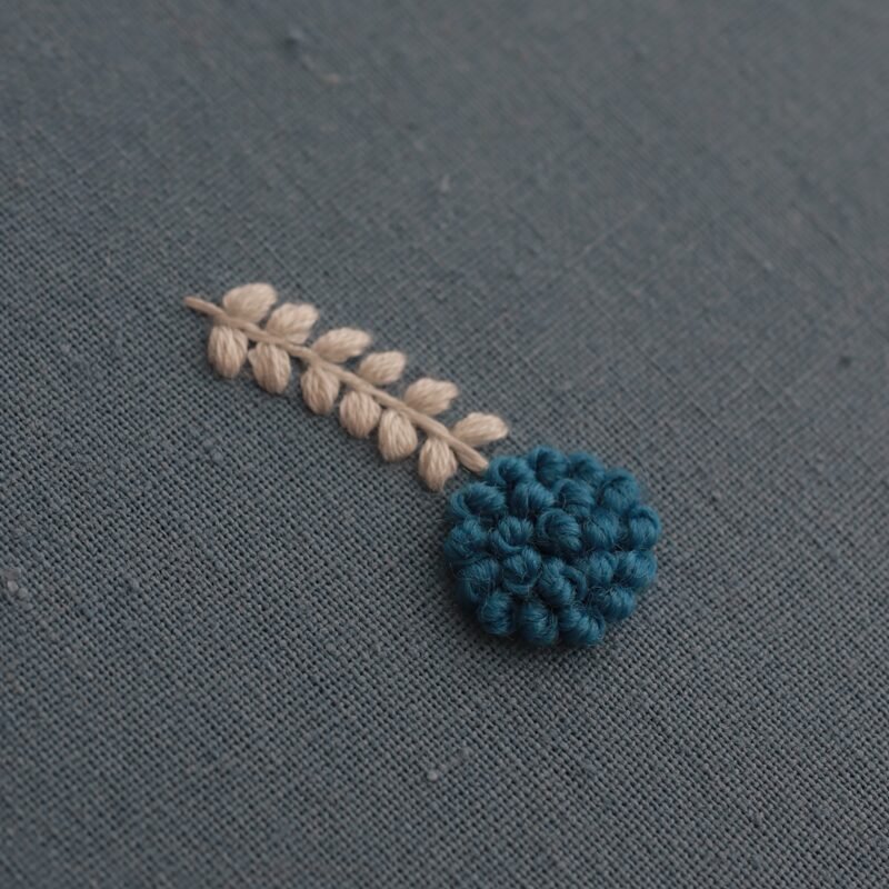Extreme close-up of a single embroidered motif by lissyluo, detailing the texture of the teal French knots and cream satin-stitched leaves.