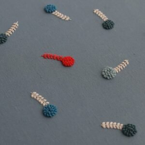Angled view of a lissyluo hand embroidery, showing scattered blue and cream botanical motifs with a single red accent on dusty blue fabric.