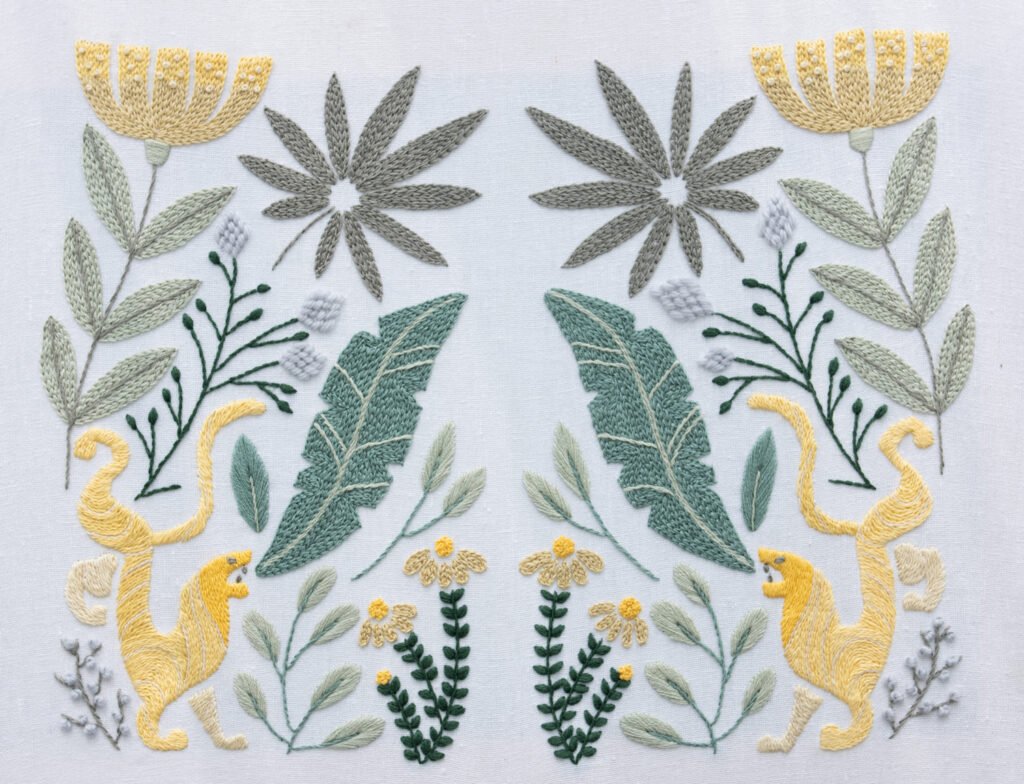 Hand embroidery titled "Rousseau's Jungle," featuring a symmetrical design of yellow leopards, tropical leaves, and stylized flowers on white fabric.
