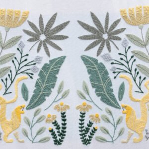 Hand embroidery titled "Rousseau's Jungle," featuring a symmetrical design of yellow leopards, tropical leaves, and stylized flowers on white fabric.