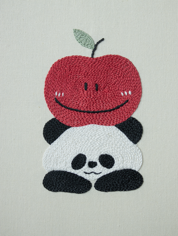 Hand-embroidered art of a smiling panda wearing a large, happy red apple like a hat, stitched on off-white fabric.