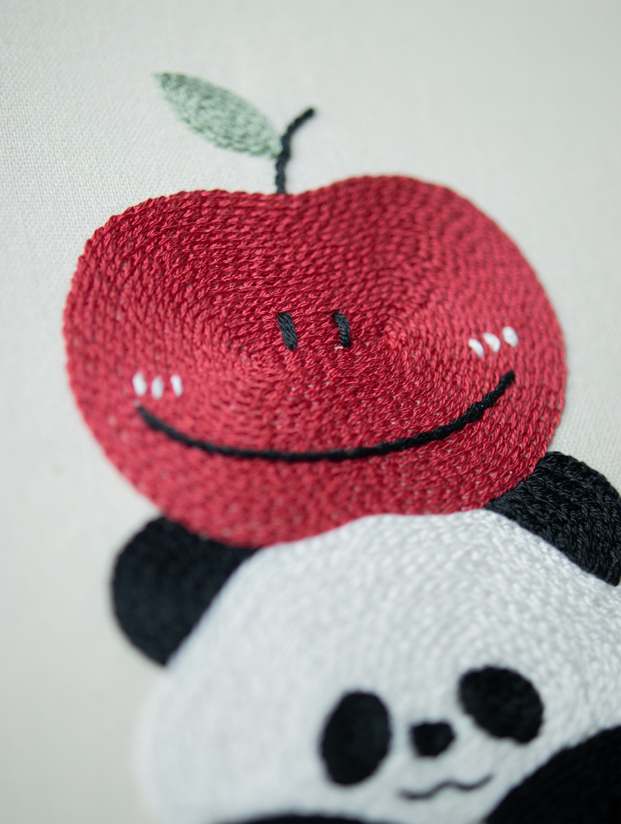 A macro photo of the Apple Panda embroidery, focusing on the smiling apple's textured red chain stitch fill and the straight line stitches of its happy face.