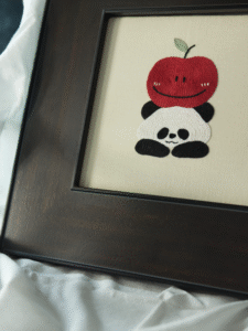 An angled view of the Apple Panda embroidery art, presented in a dark brown wooden frame and placed on soft white fabric.