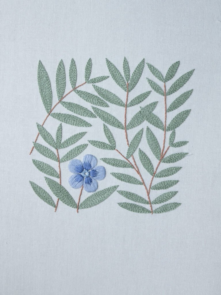 A finished hand embroidery piece featuring a modern botanical pattern with a single blue flower and sage green leaves on a light fabric background.