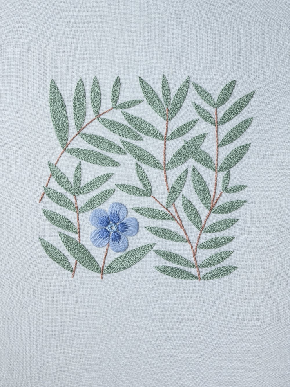Modern Botanical Hand-Embroidered Wall Art A finished hand embroidery piece featuring a modern botanical pattern with a single blue flower and sage green leaves on a light fabric background.