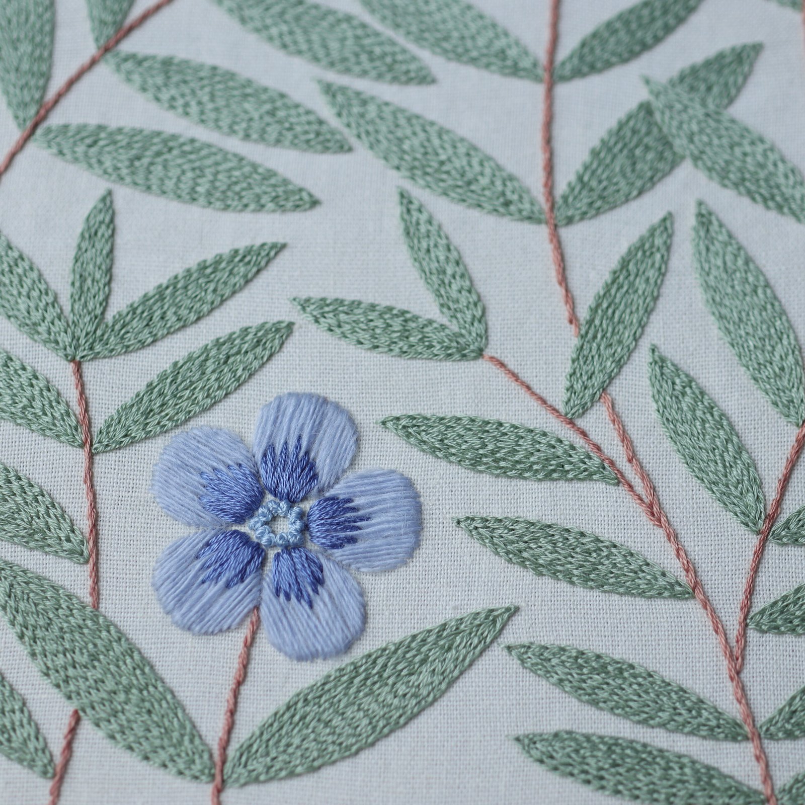 Detailed close-up of a hand-embroidered blue flower with satin stitch petals, surrounded by sage green leaves on white linen fabric.