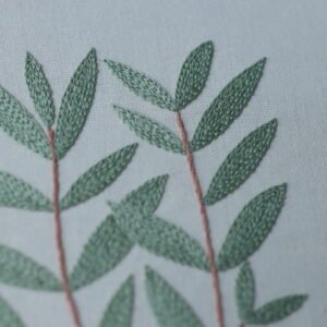 A close-up detail shot of hand-embroidered sage green leaves with satin stitch texture on a light gray fabric.