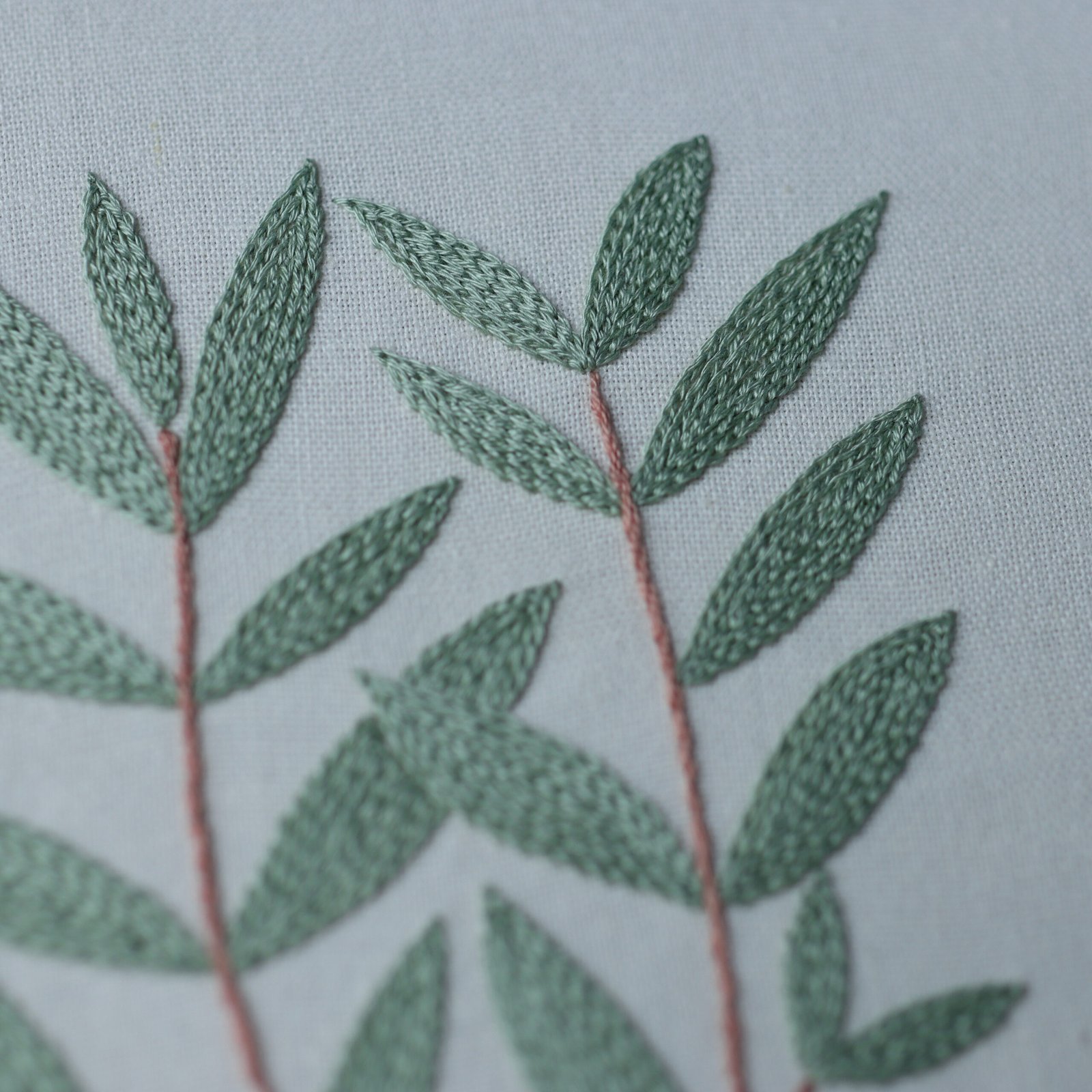 A close-up detail shot of hand-embroidered sage green leaves with satin stitch texture on a light gray fabric.