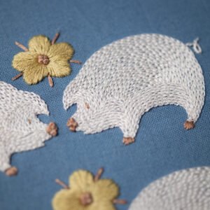 A close-up photo of two hand-embroidered white pigs touching noses, with a yellow satin-stitched flower between them on a blue fabric background.