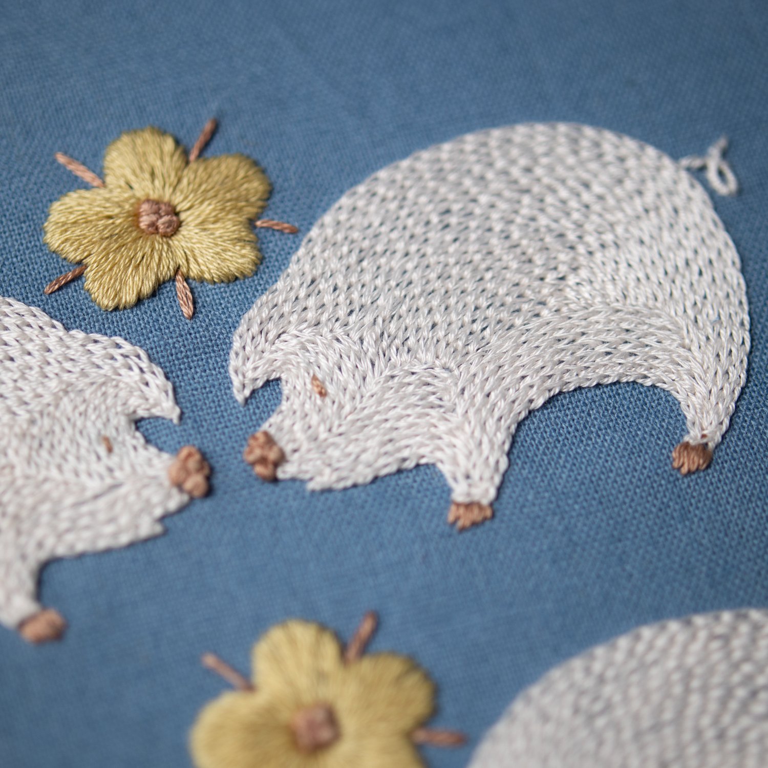 A close-up photo of two hand-embroidered white pigs touching noses, with a yellow satin-stitched flower between them on a blue fabric background.