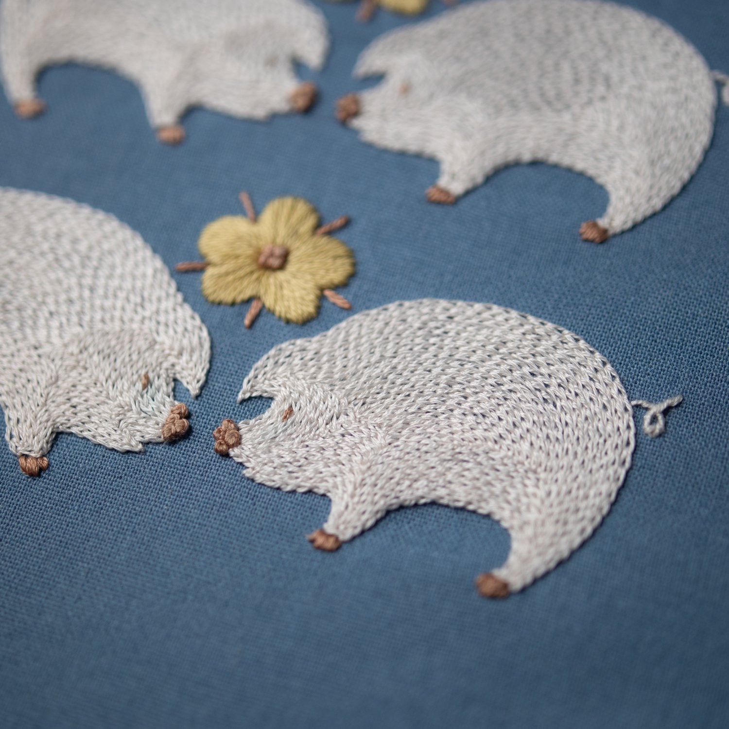 Two charming embroidered pigs, stitched in white, appear to kiss over a yellow flower on a blue canvas.