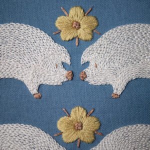 Macro view of embroidery stitch work, showing the texture of white thread on two pigs and the smooth satin stitch of a yellow flower with a French knot center.