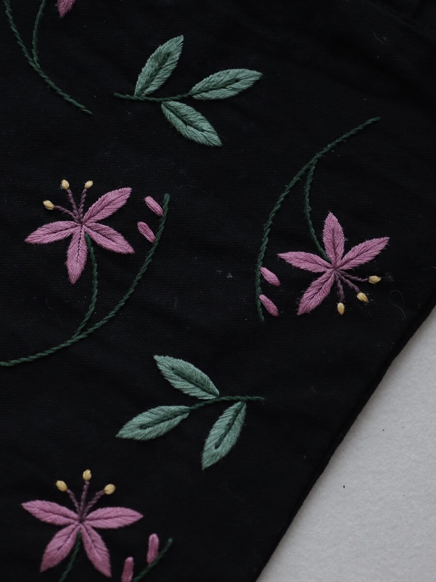 Angled close-up of the botanical embroidery, showing the texture of the pink and green thread on the black canvas fabric.