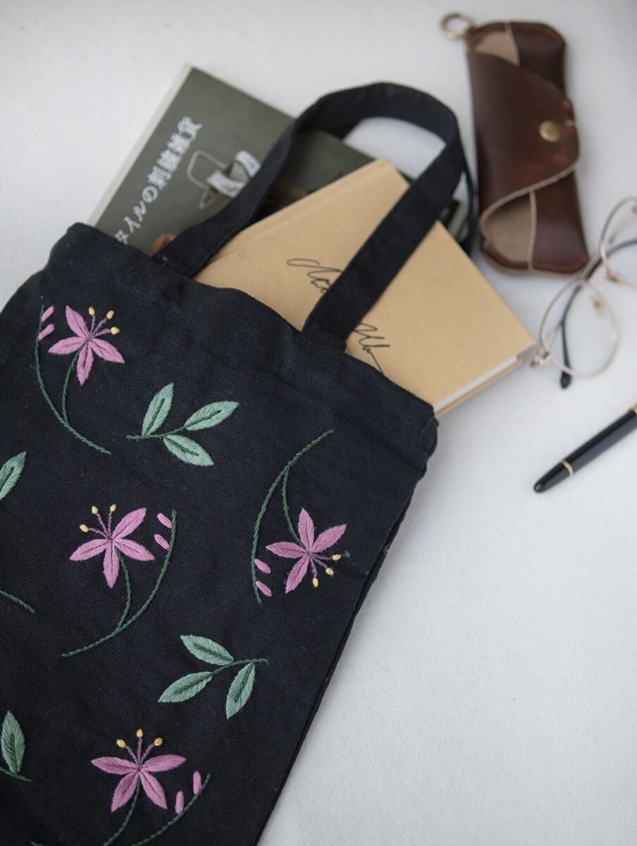 Lissyluo's hand-embroidered floral tote bag, shown holding books and a notebook, with a leather glasses case in the background.