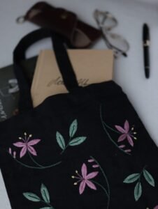 A black tote bag with hand-embroidered pink flowers, styled for daily use with books, a fountain pen, and eyeglasses.