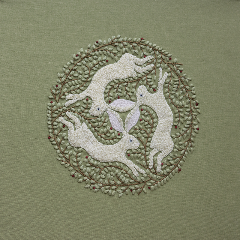 Hand-embroidered "Three Hares" motif featuring three white rabbits sharing ears in a circle, surrounded by a wreath of green leaves and red berries on a sage green fabric.