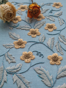 A styled photo of the hand-embroidered floral vine, with two dried roses resting on the blue linen fabric.