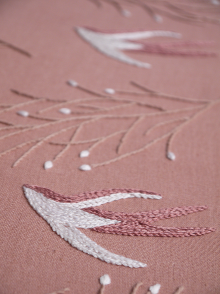 Angled close-up of a hand-embroidered swallow, showing the texture of the white and dusty rose satin stitches next to delicate branches on a mauve fabric.
