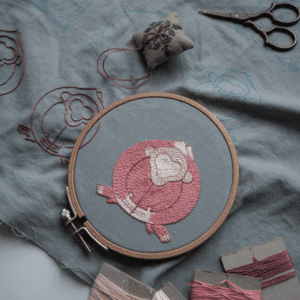 A pink monkey motif for the 'Embracing Loneliness' piece is complete in an embroidery hoop, while outlines for other figures are visible on the surrounding grey linen.