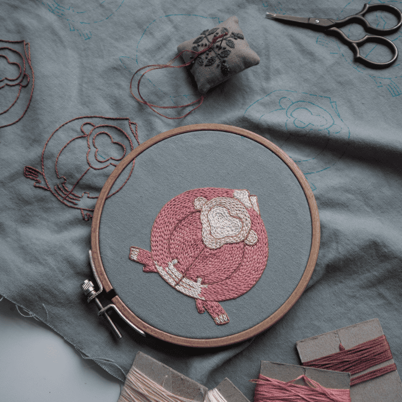 A pink monkey motif for the 'Embracing Loneliness' piece is complete in an embroidery hoop, while outlines for other figures are visible on the surrounding grey linen.