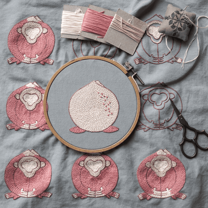 Work in progress of the 'Embracing Loneliness' hand embroidery art. The central peach figure is in a hoop, surrounded by finished monkey figures, thread spools, and scissors on grey linen.