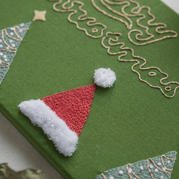 Christmas Embroidery Wall Art - Detail Shot Close-up detail of hand-embroidered Christmas wall art, showing the textured red Santa hat and the gold thread of the 'Merry Christmas' lettering.