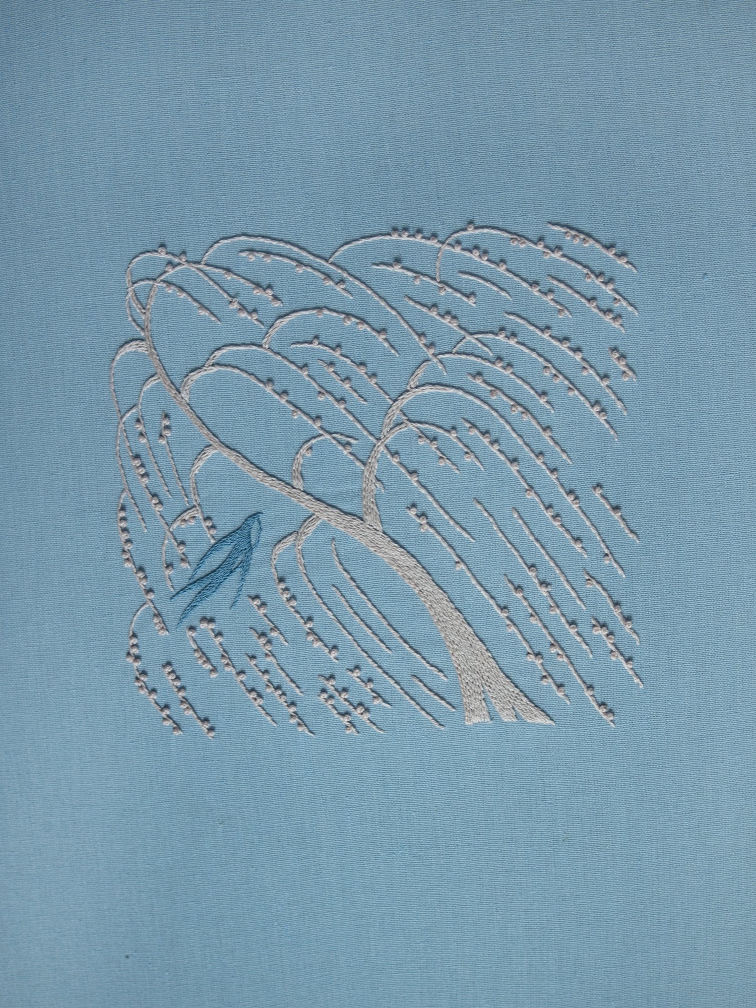 Full view of a minimalist hand-embroidery piece featuring a weeping willow tree in the wind and a small blue bird, stitched on light blue fabric.