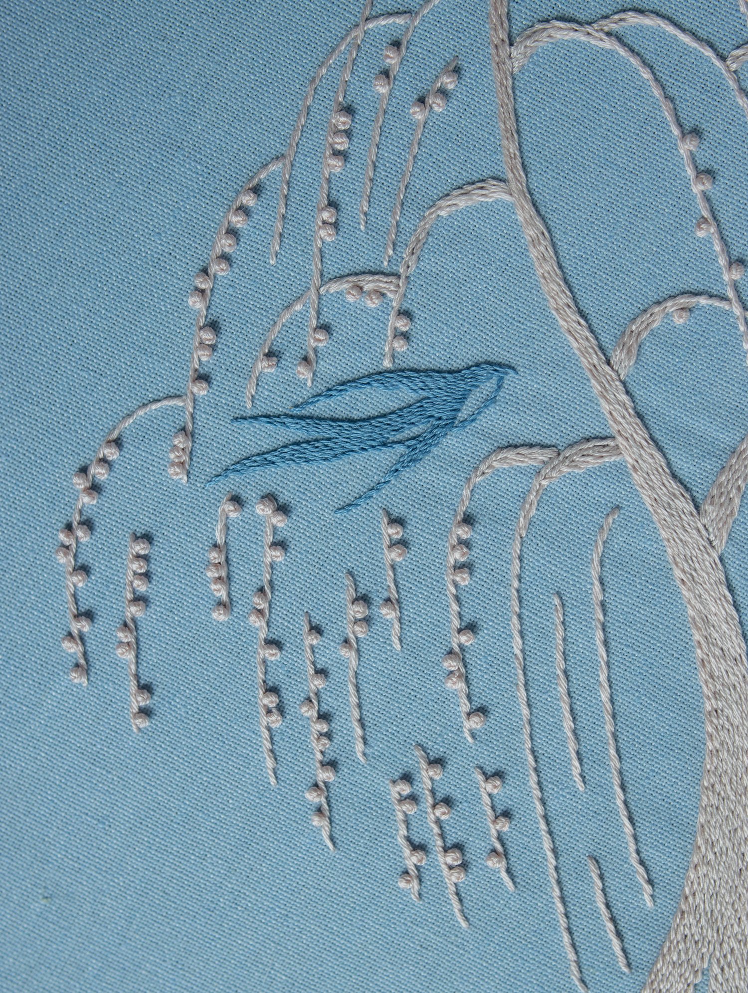 Hand-embroidered art showing a blue swallow resting on a weeping willow branch. The tree is stitched in off-white thread on a blue background.