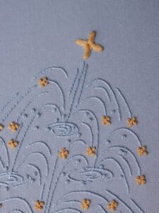Close-up of a stylized fountain hand-embroidered by lissyluo. Arcs of light blue thread spray upwards on pale blue fabric, with golden-yellow stitches like sparkles.