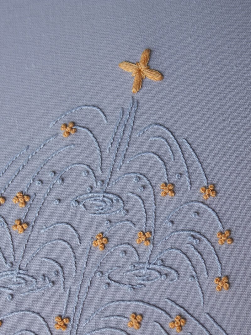 Close-up of a stylized fountain hand-embroidered by lissyluo. Arcs of light blue thread spray upwards on pale blue fabric, with golden-yellow stitches like sparkles.