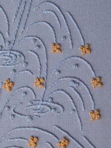 Close-up of a stylized fountain hand-embroidered by lissyluo. Arcs of light blue thread spray upwards on pale blue fabric, with golden-yellow stitches like sparkles.