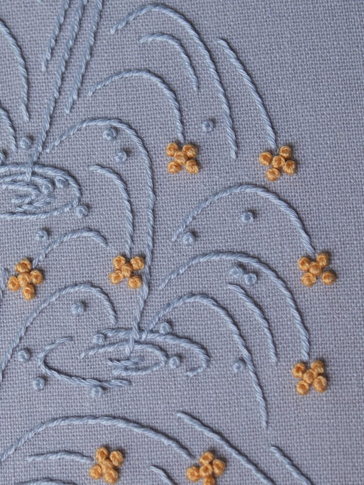 Close-up of a stylized fountain hand-embroidered by lissyluo. Arcs of light blue thread spray upwards on pale blue fabric, with golden-yellow stitches like sparkles.