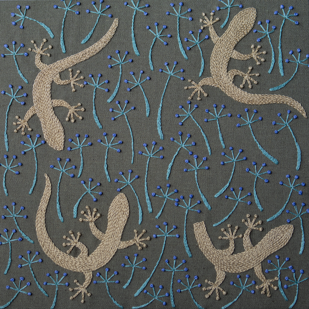 Hand-embroidered pattern of beige geckos and stylized blue flowers on dark green fabric.
