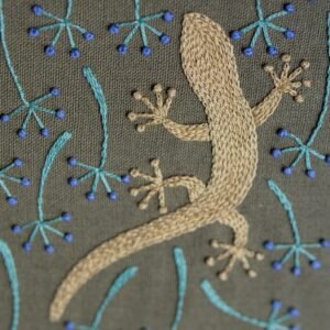 Angled close-up of a hand-embroidered gecko, showing the texture of the beige thread against the green fabric.