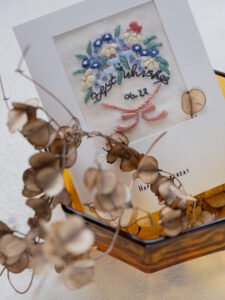Close-up of a hand-embroidered birthday card with a floral bouquet, resting in a glass dish with dried lunaria pods.