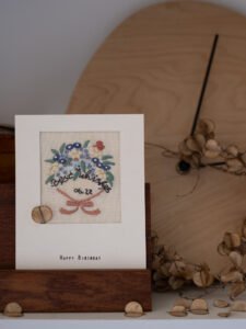 Hand-embroidered floral birthday card displayed in a wooden stand, with a minimalist wooden clock in the background.