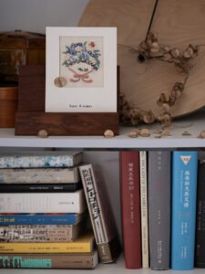 Hand-embroidered floral birthday card resting on a stack of art and literature books on a white bookshelf.