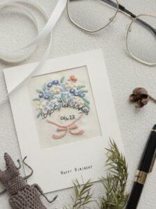 Artistic flat lay of the hand-embroidered floral card, styled with a fountain pen, eyeglasses, and a white ribbon