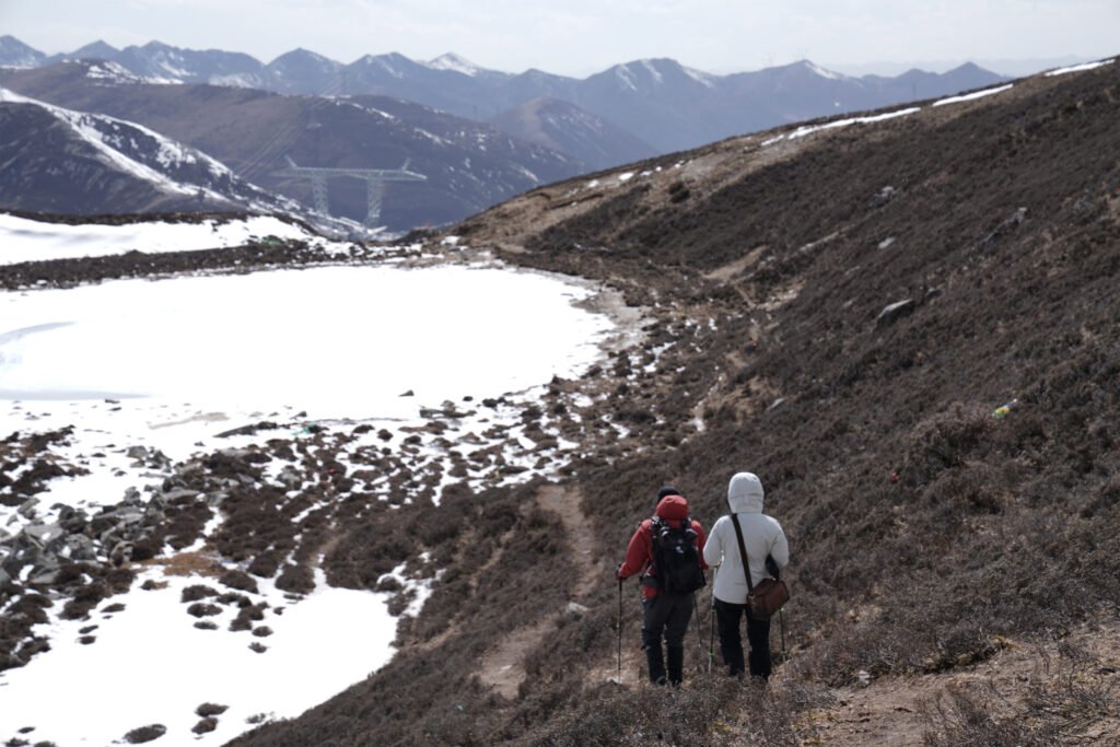 Two hikers with backpacks and poles walk along a trail on a rugged, brush-covered mountainside in Western Sichuan. To their left is a snow-covered basin, and in the distance, a vast range of snow-capped peaks rises against the sky.